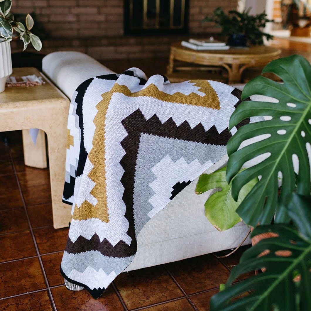 A throw blanket with a grey, white, gold, chocolate and black geometric pattern. Throw is laid out over a couch and displayed in a living room setting. The Homeland Throw is designed by Native American knitwear designer Jennifer Berg and made in New Mexico, USA.
