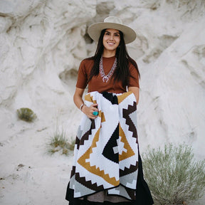 Model holds a throw blanket with a grey, white, gold, chocolate and black geometric pattern. The Homeland Throw is designed by Native American knitwear designer Jennifer Berg and made in New Mexico, USA.