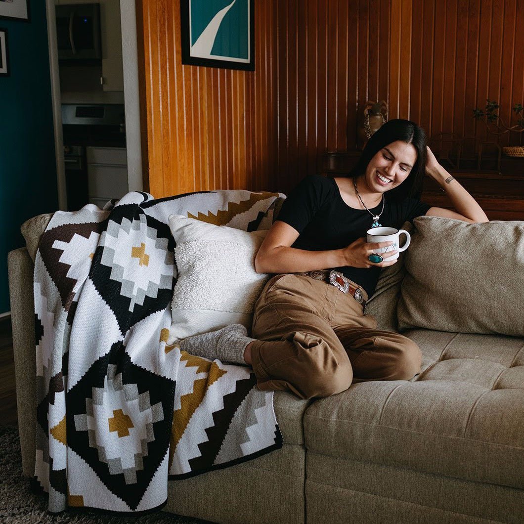 A throw blanket with a grey, white, gold, chocolate and black geometric pattern. Throw is laid out over a couch and displayed in a living room setting. The Homeland Throw is designed by Native American knitwear designer Jennifer Berg and made in New Mexico, USA.