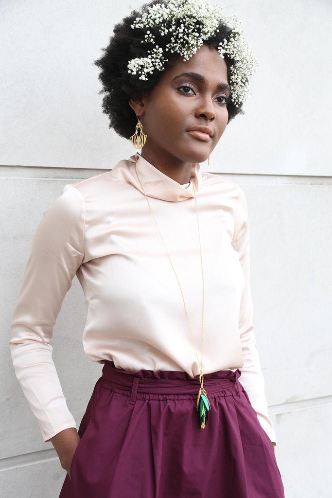 Woman with white flowers in her black hair models earrings and necklace against a white wall. The necklace is long, and measures close to the model's waist. The necklace is brass and blue and green from beetle wings. The I Can See the Light necklace is from designer Lingua Nigra.