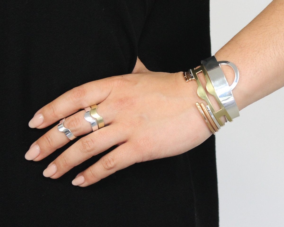 A stack of brass, 10k yellow gold, and sterling silver Mollia cuffs, a brass Makou cuff, and a silver Ela cuff, styled on a model with a black shirt and three retired betsy & iya brass and silver rings.