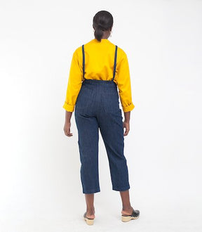 Model shows the back side of dark blue cropped overalls with thin adjustable straps and two front pockets over a long sleeve yellow shirt. The Knot Overalls in Dark Indigo are designed by Loup and made in New York City, NY.