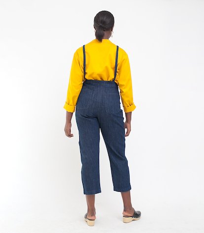 Model shows the back side of dark blue cropped overalls with thin adjustable straps and two front pockets over a long sleeve yellow shirt. The Knot Overalls in Dark Indigo are designed by Loup and made in New York City, NY.