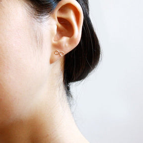 A model wears a gold tone stud earring in the infinity symbol. The Infinity Stud Earring is designed by Hooks and Luxe and handcrafted in Jackson Heights, NY.