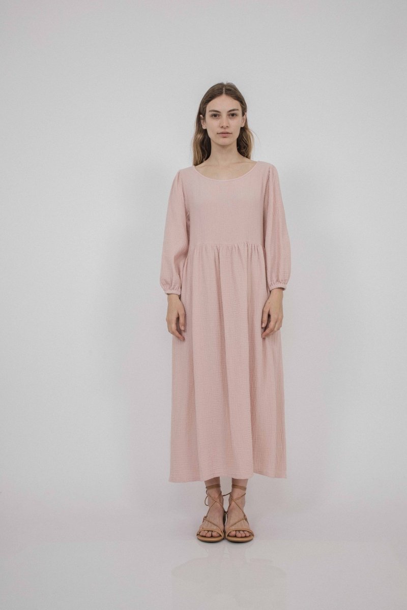 A model wears a 3/4 sleeve dress with ruching details. The dress has elastic armhole and side pockets in a light pink color. The Juniper Dress in Blush is designed by Corinne Collection in Los Angeles, CA.