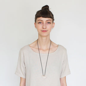 The betsy & iya black Japanese cotton rope and cast bronze semicircle Kanssa necklace, pictured on a model wearing a pale gray tunic.