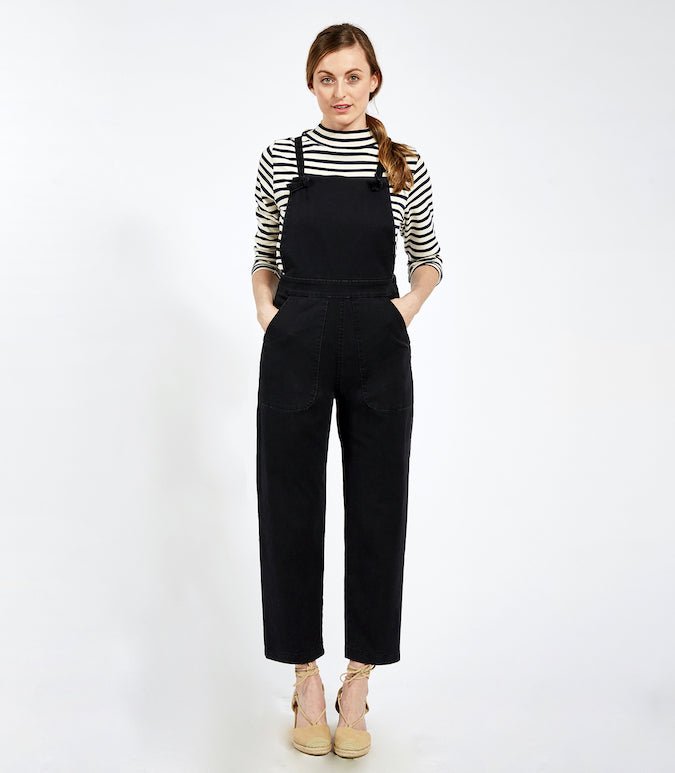 Model wears cropped black overalls with thin adjustable straps and two front pockets over a black and white striped turtleneck shirt. The Knot Overalls in Black are designed by Loup and Made in New York City, NY.