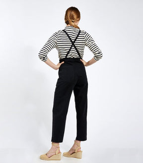 Model shows the back side of  cropped black overalls with thin adjustable straps and two back pockets over a black and white striped turtleneck shirt. Overalls are backless with adjustable straps criss-crossed. The Knot Overalls in Black are designed by Loup and Made in New York City, NY.