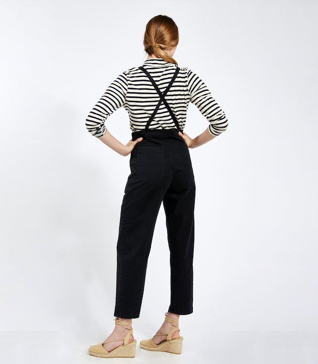 Model shows the back side of  cropped black overalls with thin adjustable straps and two back pockets over a black and white striped turtleneck shirt. Overalls are backless with adjustable straps criss-crossed. The Knot Overalls in Black are designed by Loup and Made in New York City, NY.