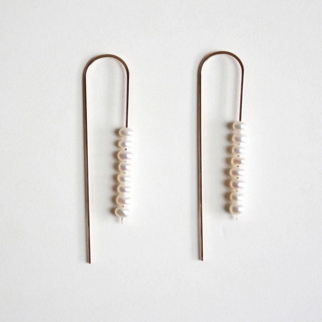 A gold tone u-shaped earring with multiple freshwater pearls stacked on one side. The Long arc Threader Earrings with Multiple Small Pearls are designed by Hooks and Luxe and handcrafted in Jackson Heights, NY.