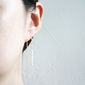 A model wears a gold tone u-shaped earring with multiple freshwater pearls stacked on one side. The Long arc Threader Earrings with Multiple Small Pearls are designed by Hooks and Luxe and handcrafted in Jackson Heights, NY.