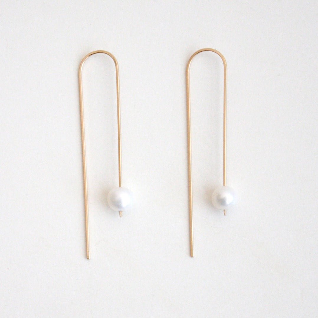 A long gold tone u-shaped earring with a single large pearl. The Long Arc Threader Earrings with Large Pearl is designed by Hooks and Luxe and handcrafted in Jackson Heights, NY.