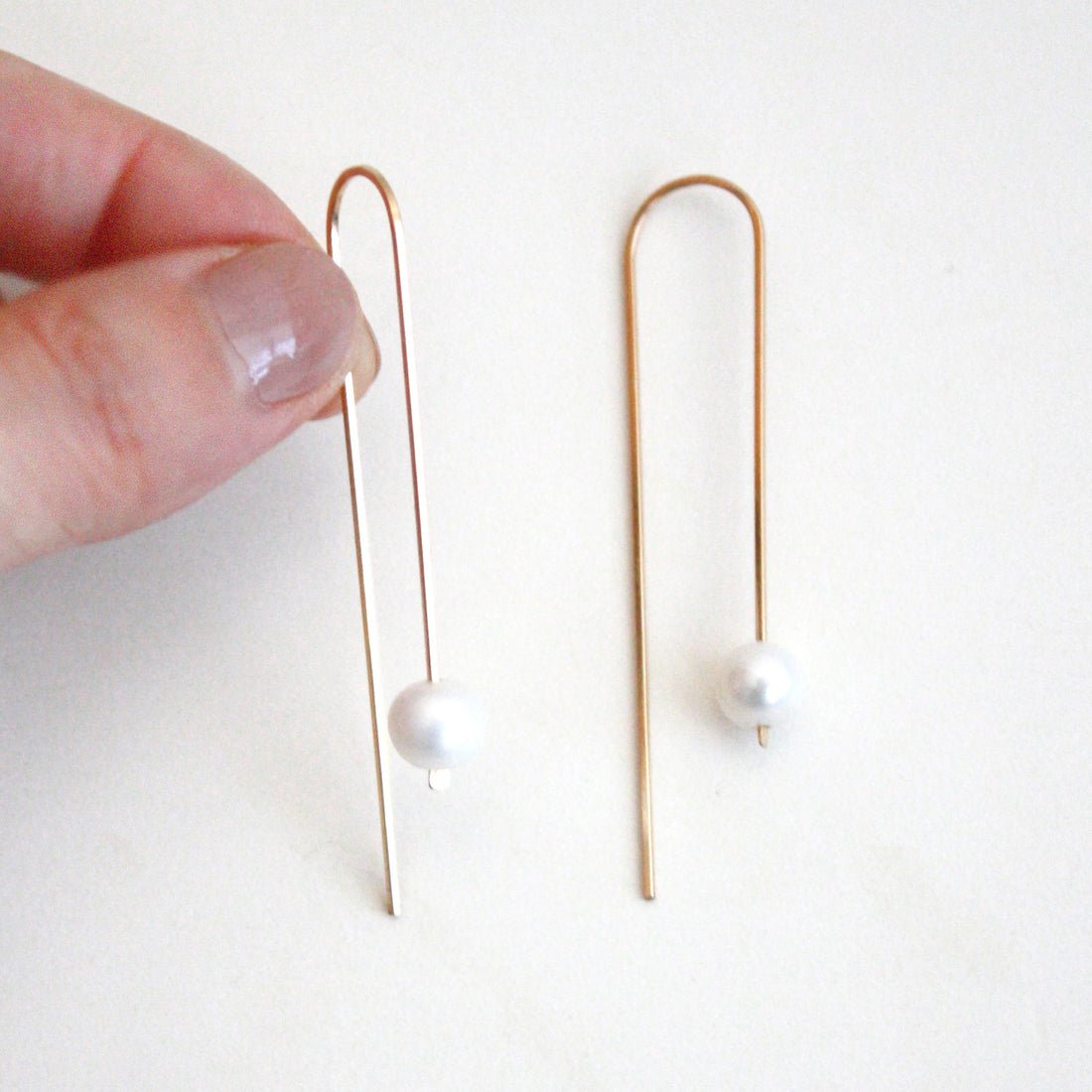 A long gold tone u-shaped earring with a single large pearl. The Long Arc Threader Earrings with Large Pearl is designed by Hooks and Luxe and handcrafted in Jackson Heights, NY.