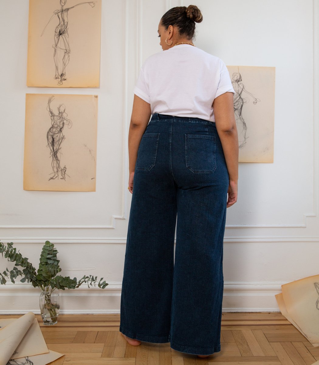A model shows the backside of long wide legged denim pants with large front pockets. The Long Sabrina Jeans in Dark Indigo are designed by Loup and made in New York City, USA.