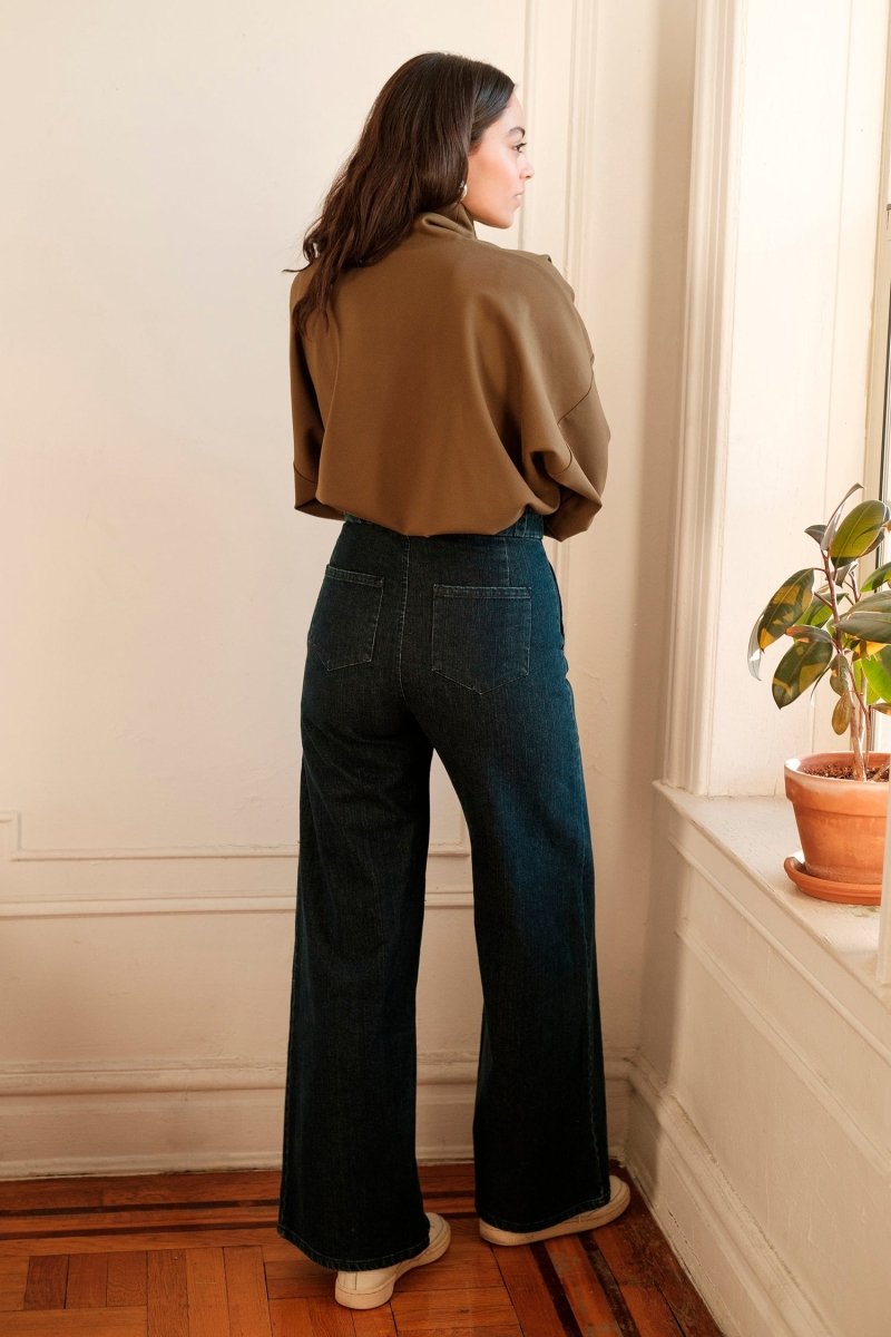 A model shows the backside of long wide legged denim pants with large front pockets. The Long Sabrina Jeans in Dark Indigo are designed by Loup and made in New York City, USA.