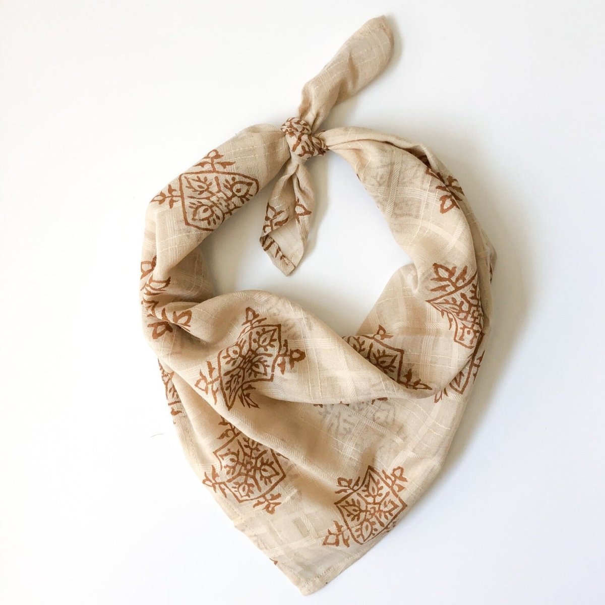 A golden brown and cream patterned bandana, folded over and tied in a knot. Block printed by hand, the Gold Compass Bandana from Maelu is designed in Portland, Oregon and handmade in India.