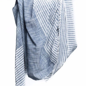 Blue scarf with white stripes and cotton fringe hangs in the air. The Malabar Natural Khadi Scarf in Blue is from Bloom & Give and hand-loomed in South India.