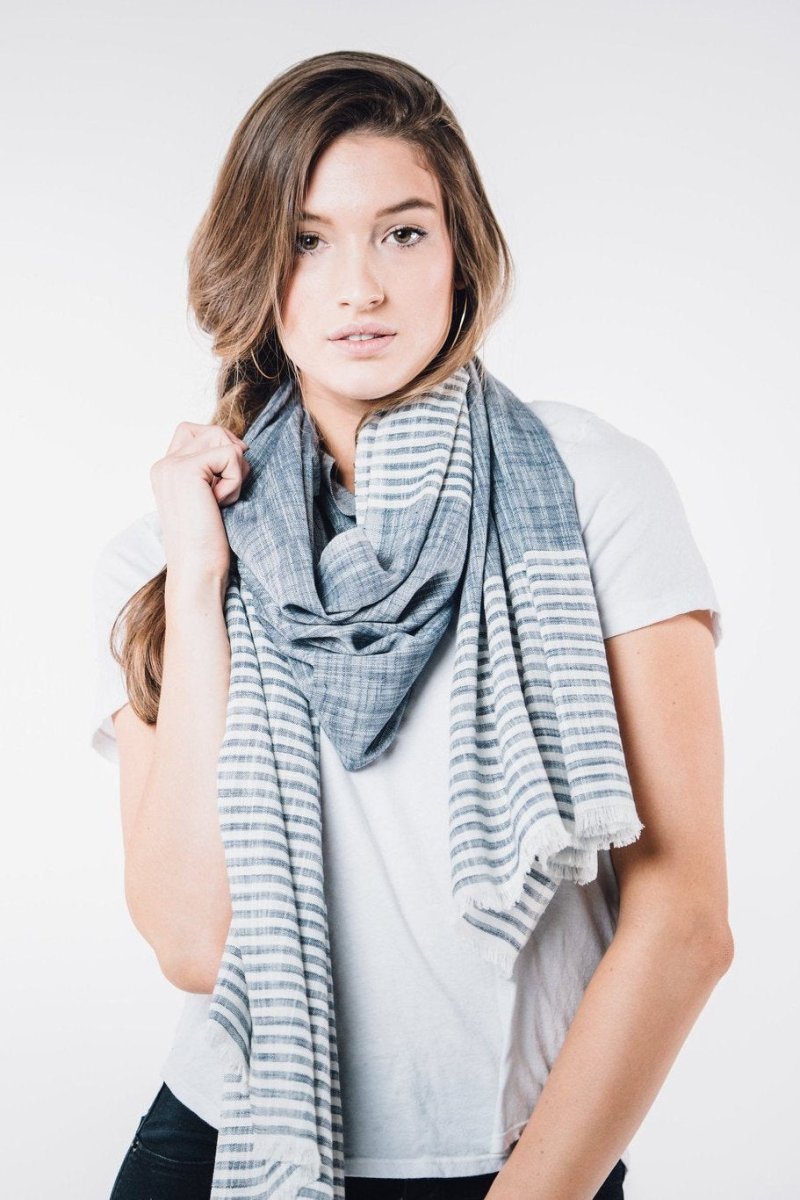 Blue scarf with white stripes and cotton fringe worn on model with light brown hair wearing a white t-shirt. The Malabar Natural Khadi Scarf in Blue is from Bloom & Give and hand-loomed in South India.