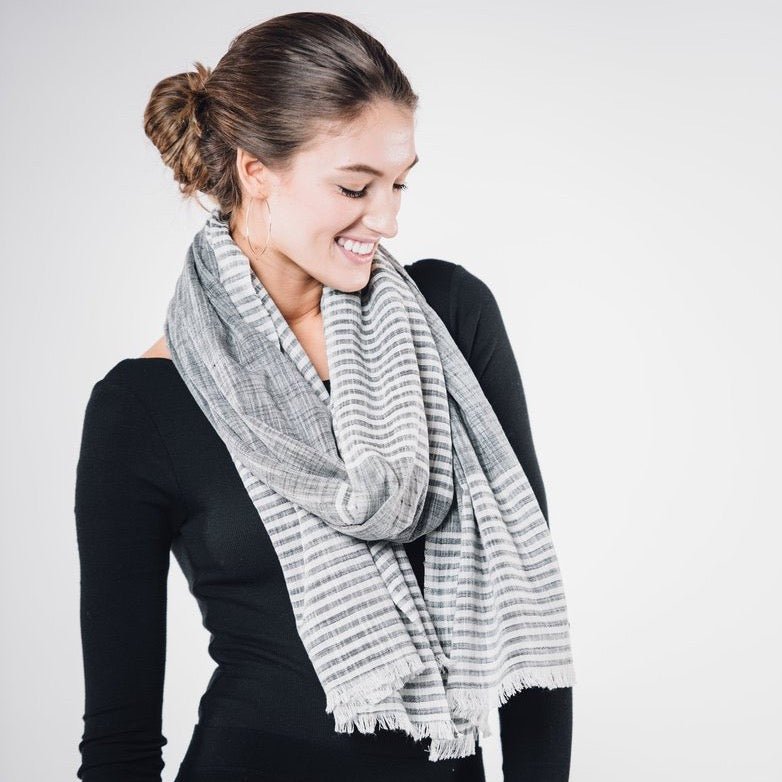 Grey scarf with white stripes and cotton fringe worn on model with brown hair and a long sleeve black shirt. The Malabar Natural Khadi Scarf in Grey is from Bloom & Give and hand-loomed in South India.