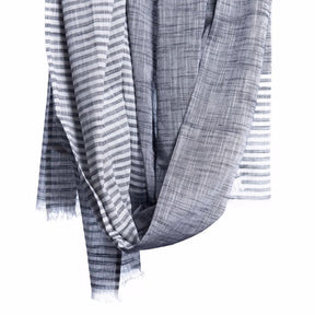 Grey scarf with white stripes and cotton fringe detail hangs in the air. The Malabar Natural Khadi scarf in Grey is from Bloom & Give and hand-loomed in South India.