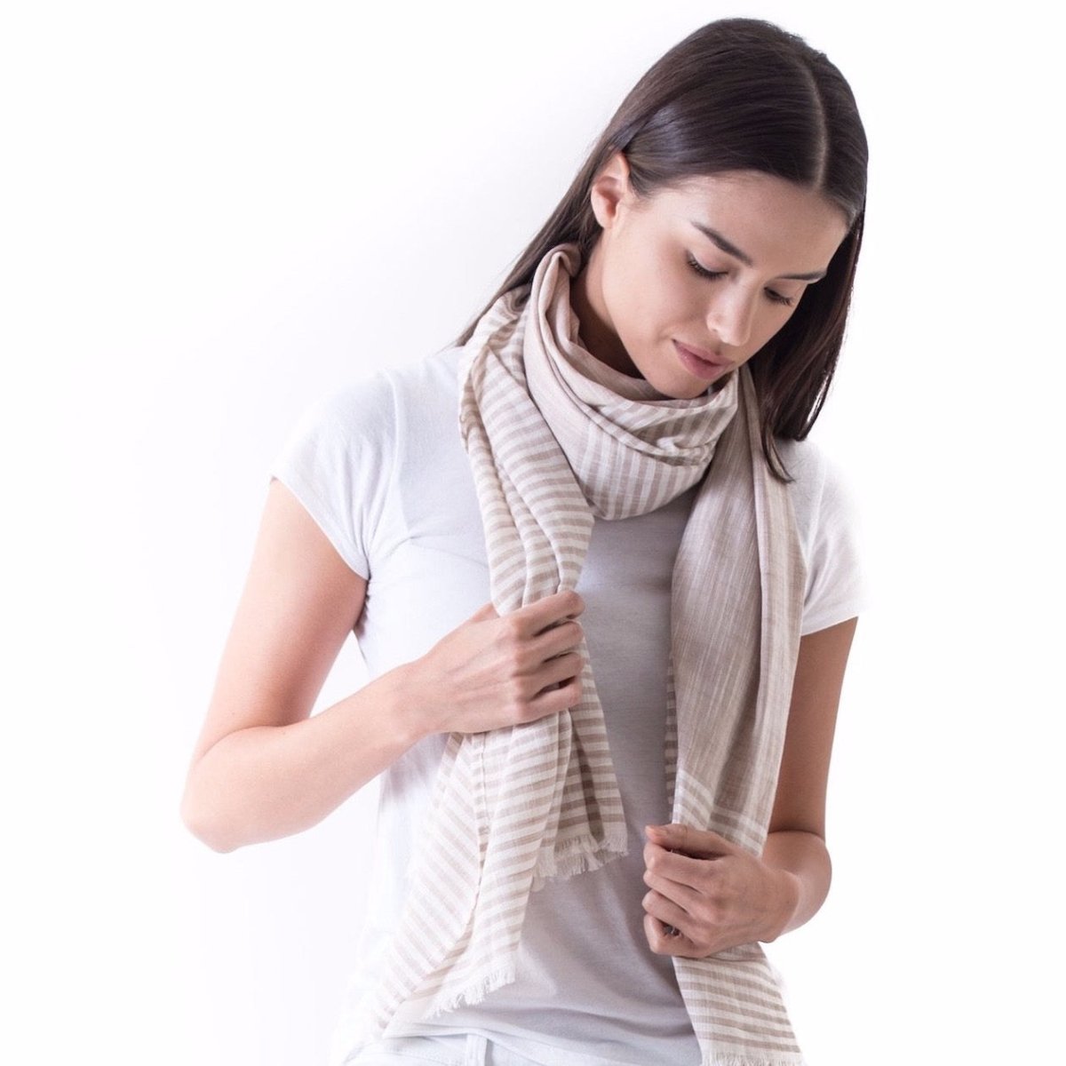 The Malabar Natural Khadi Scarf worn on a model with dark brown hair wearing a white t-shirt. This natural cotton scarf with white stripes and fringe is from Bloom & Give and hand-loomed in South India.