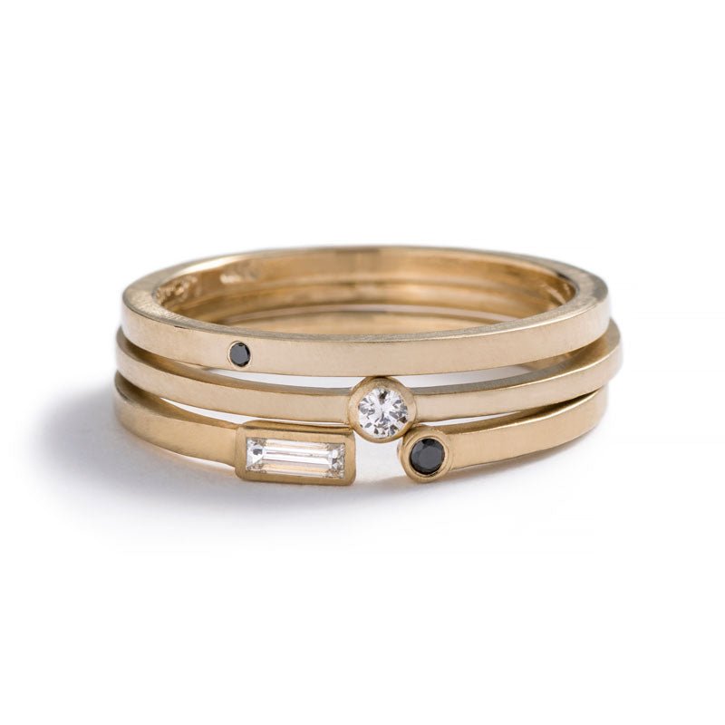 A stack of three betsy & iya 14k yellow gold rings: Vis Ring with a round, flush-set black diamond, Robur Ring with a round, bezel-set white diamond, and Manus Ring with a white diamond baguette and round black diamond. Hand-crafted in Portland, Oregon.