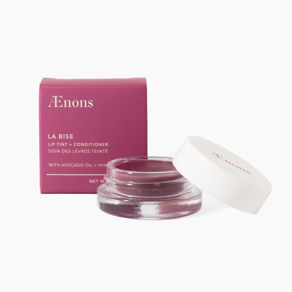 A square magenta package stands to the left of a small cylindrical container of berry colored lip tint. The Lip Tint & Conditioner in Melrose is crafted by Aenons and made in Los Angeles, CA.