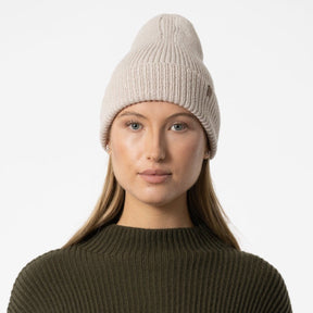 A model wears a cream colored knitted and cuffed hat with a decrease detail on top. The Merino Rib Hat in Almond White is designed by Dinadi and hand knitted in Kathmandu, Nepal.