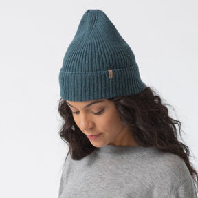 A model wears a cuffed blue/green knitted hat with a decrease detail on top. The Merino Rib Hat in Dragonfly Green is designed by Dinadi and hand knitted in Kathmandu, Nepal.