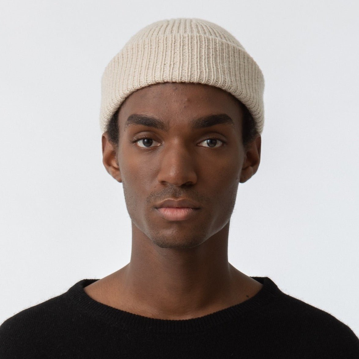 A model wears a rolled up cream colored knitted and cuffed hat with a decrease detail on top. The Merino Rib Hat in Almond White is designed by Dinadi and hand knitted in Kathmandu, Nepal.