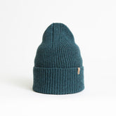 A blue/green knitted and cuffed hat with a decrease detail on top. The Merino Rib Hat in Dragonfly Green is designed by Dinadi and hand knitted in Kathmandu, Nepal.