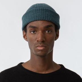 A model wears a rolled and cuffed blue/green knitted hat with a decrease detail on top. The Merino Rib Hat in Dragonfly Green is designed by Dinadi and hand knitted in Kathmandu, Nepal.