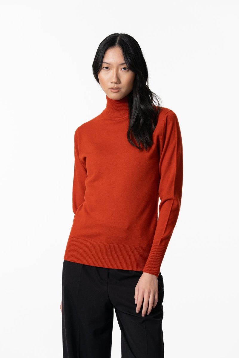 A model wears a long sleeve knitted mock neck turtleneck in a bright orange. The Merino Turtleneck in Burnt Orange is designed by Dinadi and hand knitted in Kathmandu, Nepal.