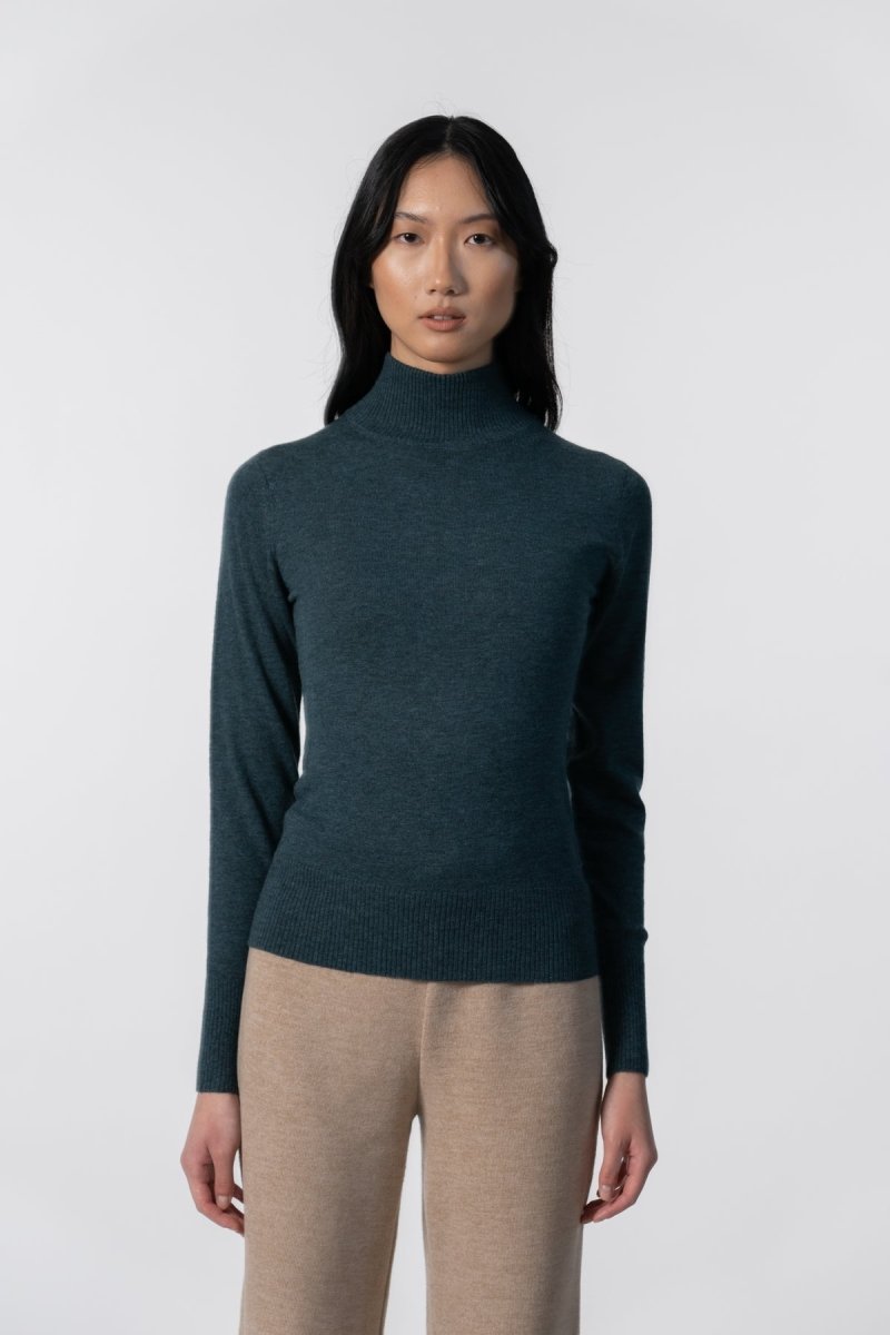 A model wears a long sleeve knitted mock neck turtleneck in a dark teal. The Merino Turtleneck in Dragonfly Green is designed by Dinadi and hand knitted in Kathmandu, Nepal.