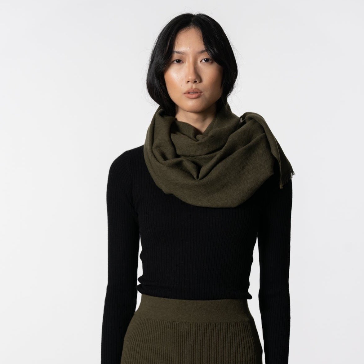 A model wears a dark green scarf made of 100% wool. The Merino woven Scarf in Olive Green is designed by Dinadi and handmade in Nepal.