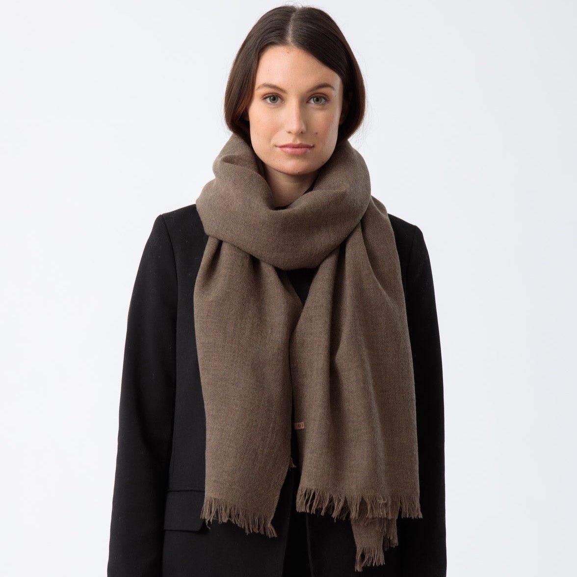 Model wears a brown scarf loosely wrapped around the neck and hangs down the front. The Merino Woven Scarf in Nutmeg Brown is from designed by Dinadi and handcrafted in Nepal.