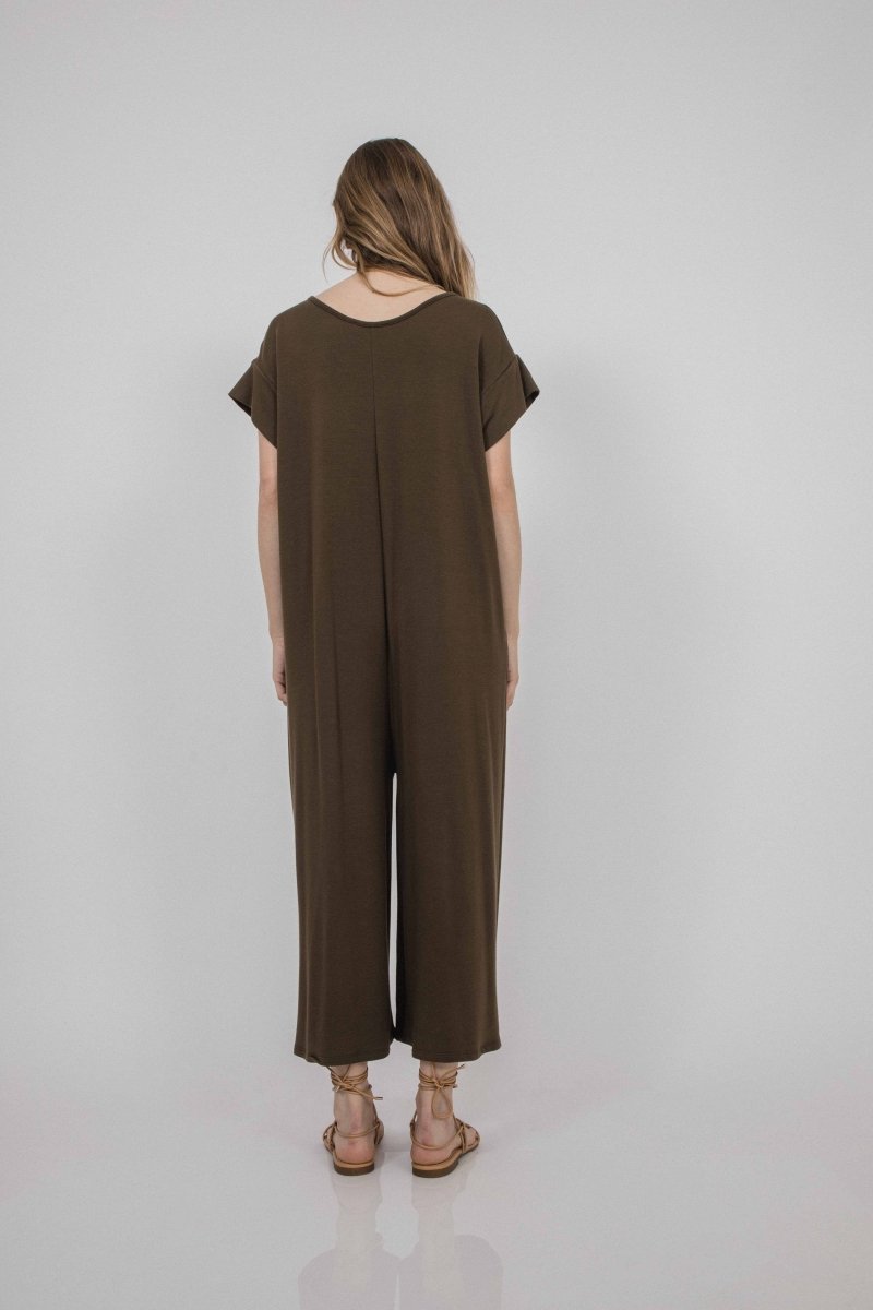 A model shows the backside of a crewneck short sleeve jumpsuit in a rich brown. It has a cuffed sleeve detail and is easy to take on and off. The Miriam Jumpsuit in mocha is designed and made by Corinne Collection in Los Angeles, CA.