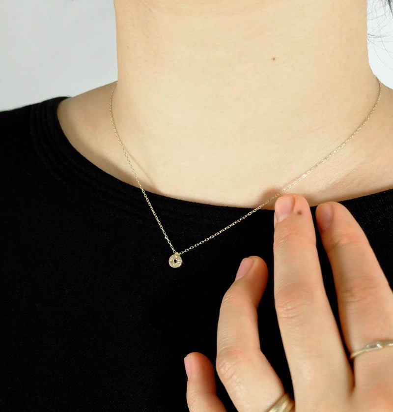 Tiny, circular pendant of 14k yellow gold, with subtle engraved details around a small black diamond, on a 14k gold chain; styled on a model with a black shirt and betsy & iya white diamond Nitor ring.