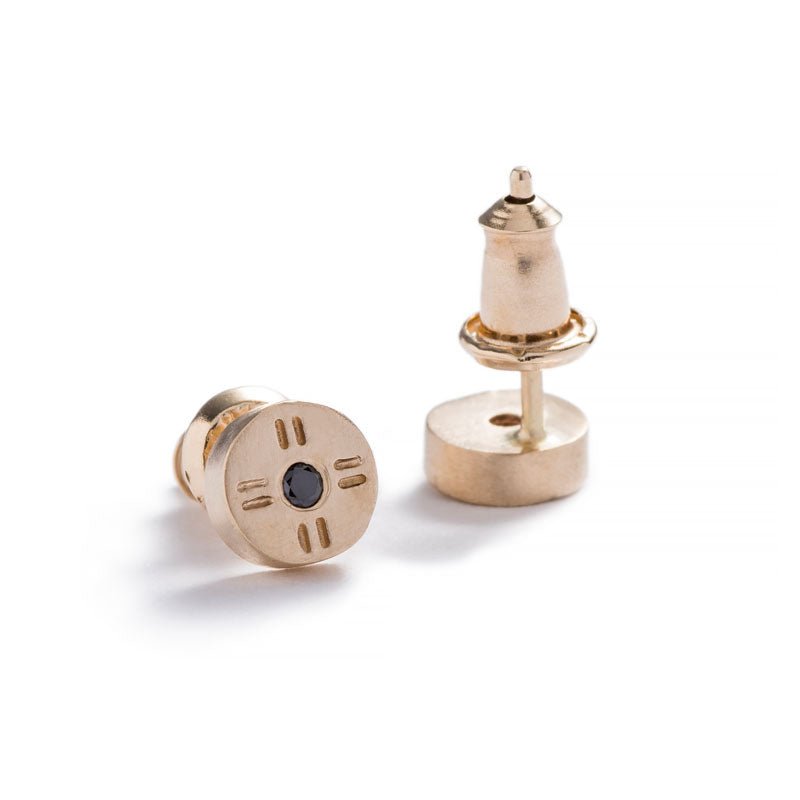 Tiny, circular stud earrings of 14k yellow gold, with subtle engraved details around a round, black diamond, and 14k gold ear nuts. Hand-crafted in Portland, Oregon.