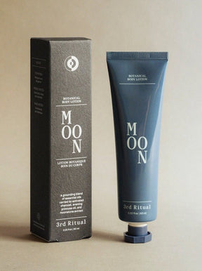 A blue tube of lotion stands next to a grey rectangular box. The Moon Botanical Body Lotion is developed and formulated by 3rd Ritual in New York City, NY.