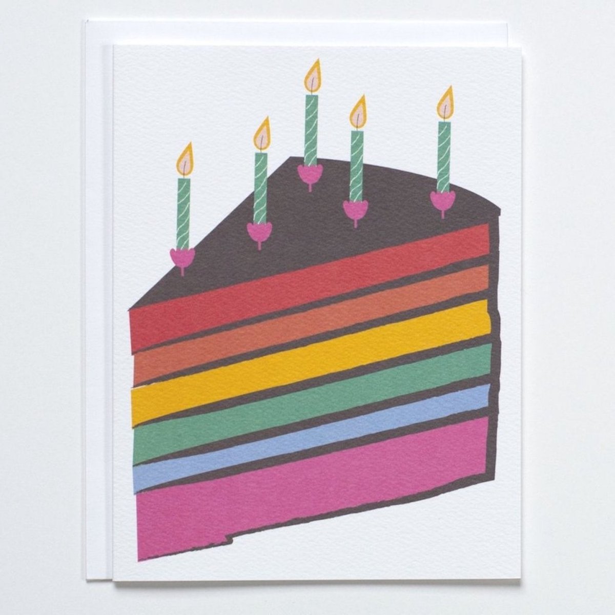 White card with a large illustration of a birthday cake slice in the colors of the rainbow. Cake slice is topped with green and pink candles with yellow and orange flames. Made with recycled paper by Banquet Atelier in Vancouver, British Columbia, Canada.