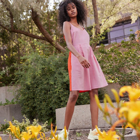 A model wears a short dress with straps and front pleats. The front portion is in a soft pink while the back is in a bright poppy color. The Nemesis Dress in Bubblegum & Rouge is designed by Eve Gravel and made in Montreal, Canada.