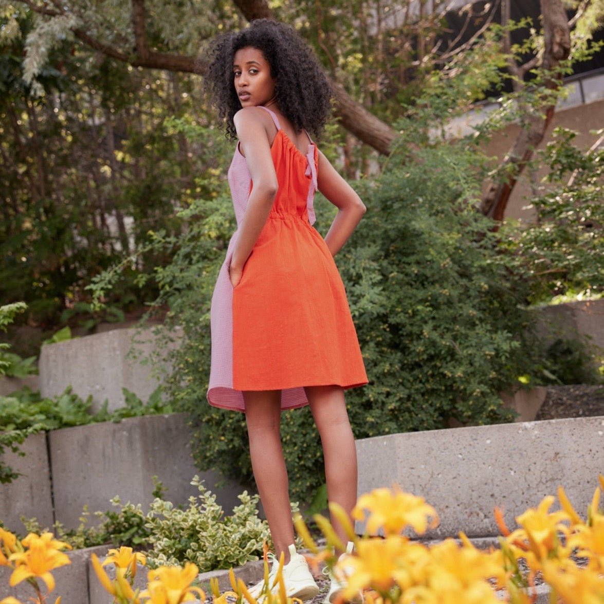 A model shows the back side of a short dress with pockets and straps that tie in the back. The Nemesis Dress in Bubblegum & Rouge is designed by Eve Gravel and made in Montreal, Canada.