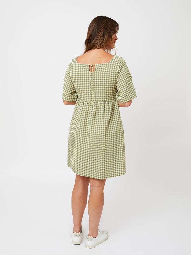 A model shows the backside of a short sleeve midi length dress with a square neckline and keyhole tieback. The fabric is in a sage green and white gingham pattern. The Nico Dress in Sage Gingham is designed by Mata Traders and made in Nepal.
