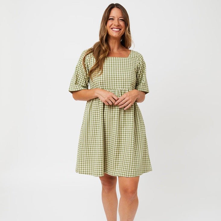 A model wears a short sleeve midi length dress with a square neckline. The fabric is in a sage green and white gingham pattern. The Nico Dress in Sage Gingham is designed by Mata Traders and made in Nepal.