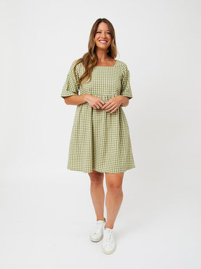 A model wears a short sleeve midi length dress with a square neckline. The fabric is in a sage green and white gingham pattern. The Nico Dress in Sage Gingham is designed by Mata Traders and made in Nepal.
