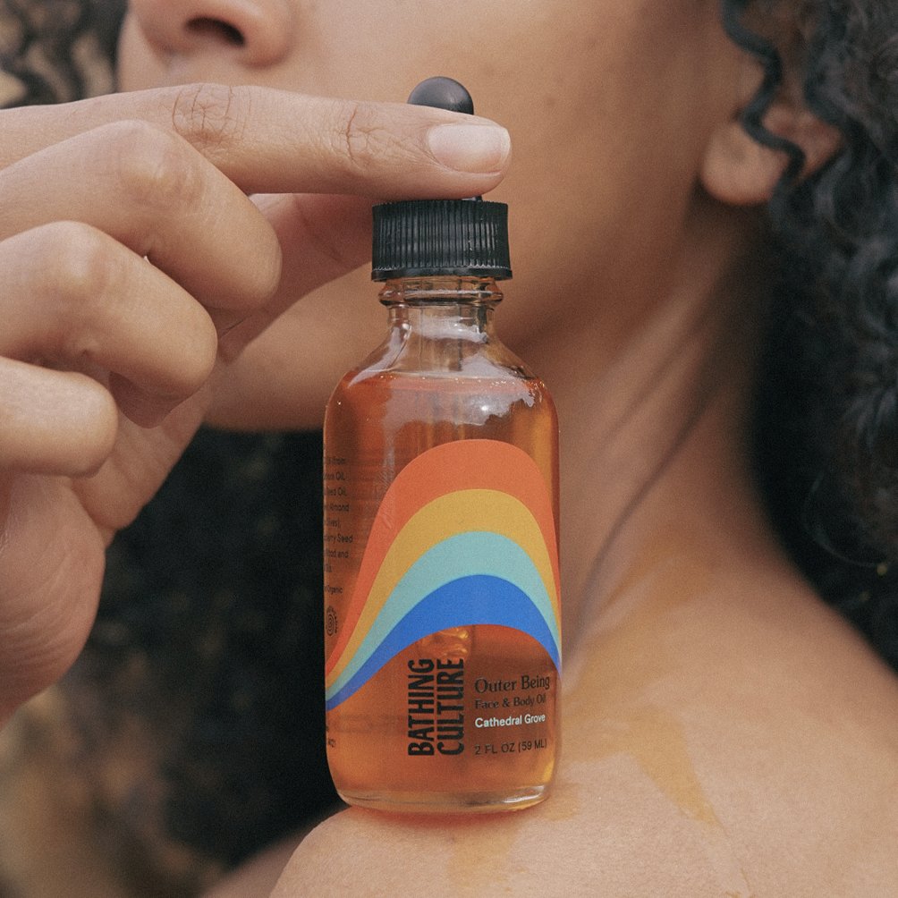 A clear glass bottle with a black dropper top holds a face and body oil. The Outer Being Face & Body oil is handcrafted by Bathing Culture in San Francisco, California.