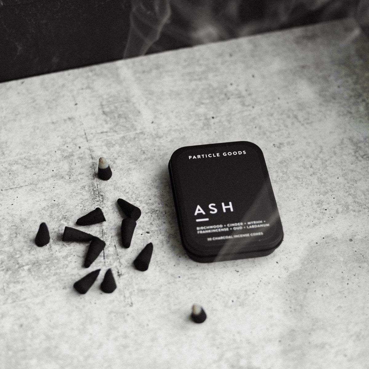 Rectangular matte black tin set next to burning  incense cones in the scent Ash. Made by Particle Goods in Seattle, WA.