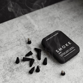 Rectangular matte black tin set next to burning incense cones in the scent Smoke. Made by Particle Goods in Seattle, WA.