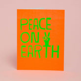 A bright orange card with an illustration of a hand making a peace sign reads: "PEACE ON EARTH." Designed by Ashkahn and printed in Portland, Oregon.